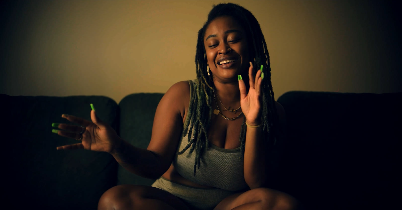 Emani laughing with her hands raised, seated on a dark sofa in warm light